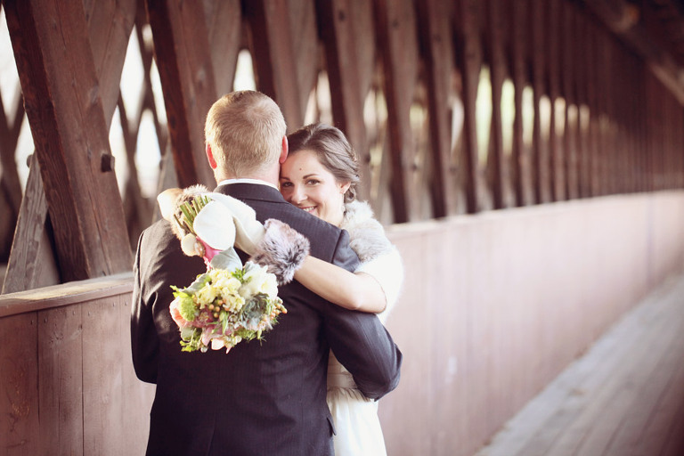 A bride smiles at the camera while hugging her husband on a covered bridge. 