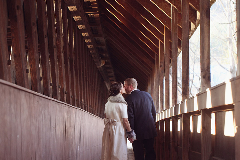 A bride and groom kiss while walking along the side of a covered bridge in Woodstock Vermont. 