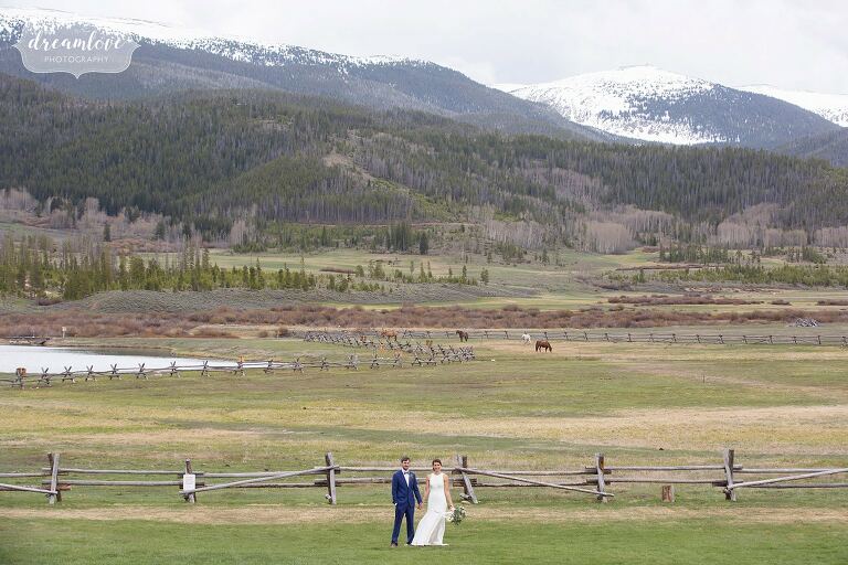 Bride and groom stand in field at Devil's Thumb Ranch surrounded by mountains and horses.
