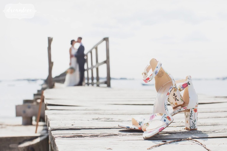 Unique wedding photo of the bride's shoes in the foreground with bride and groom on the pier in Manchester, MA.