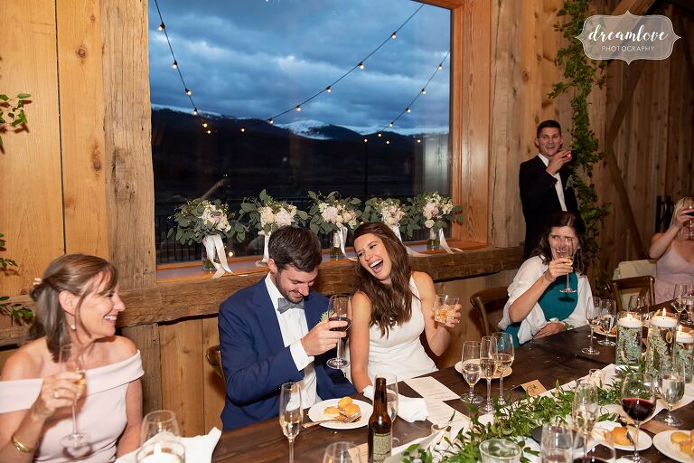 Bride and groom laugh during toasts with scenic mountain backdrop.