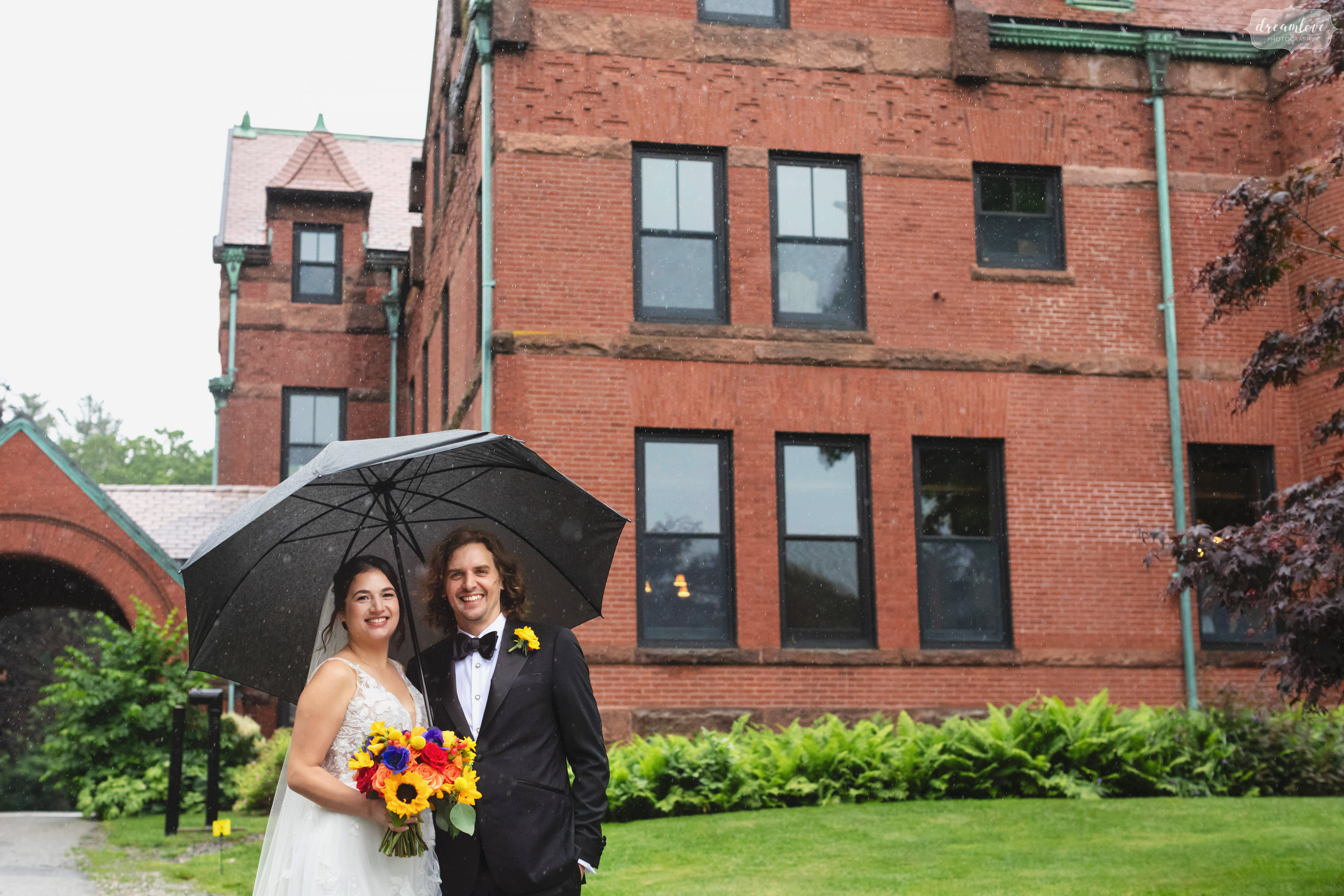Bride and groom stand under an umbrella in front of the Stevens Estate on Osgood Hill.