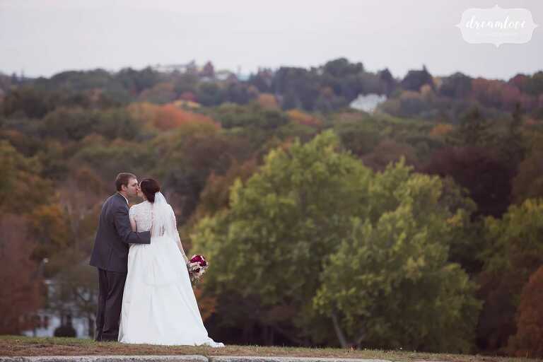 The bride and groom kiss on the hilltop with October fall foliage behind them at the Larz Anderson Park venue in Boston.