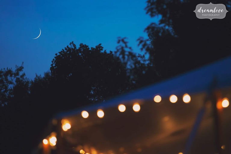 Crescent moon over cafe lights in reception tent with blue twilight.