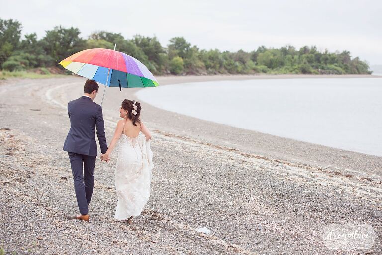 The bride and groom walk along the shore on Thompson Island with a rainbow umbrella.