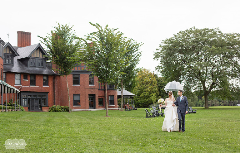 Bride and groom walk with umbrellas in front of Shelburne Farms Inn.
