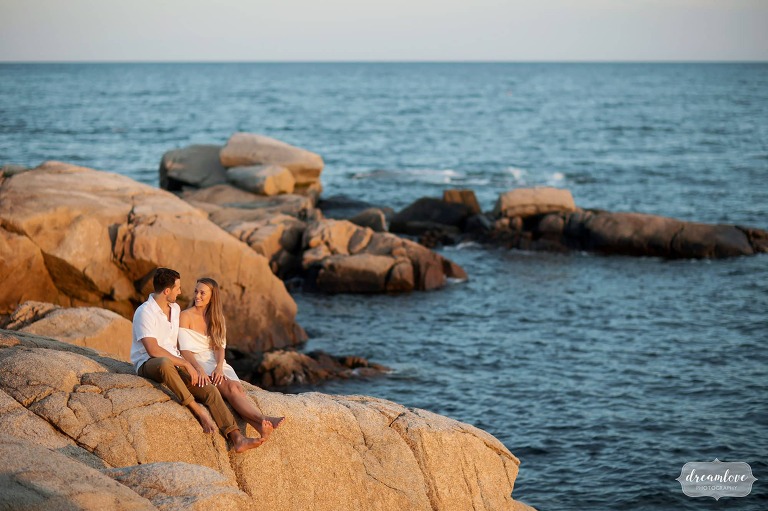Couple sits on rocks at sunset on the ocean for engagement photos.
