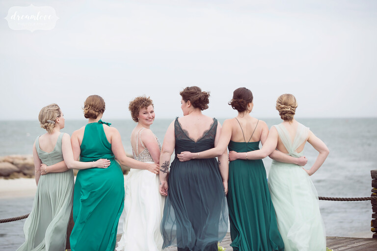 Simple photo of the bride looking back at the camera with her arms around her bridesmaids looking out at the ocean. Love their various green and blue dresses for this coastal wedding.