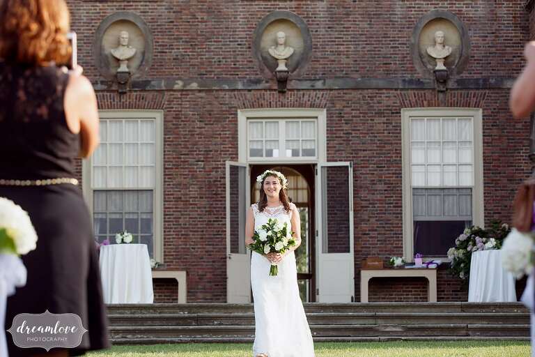 Bride walks out of the Crane Estate Castle Hill house down the aisle of her outdoor wedding ceremony.