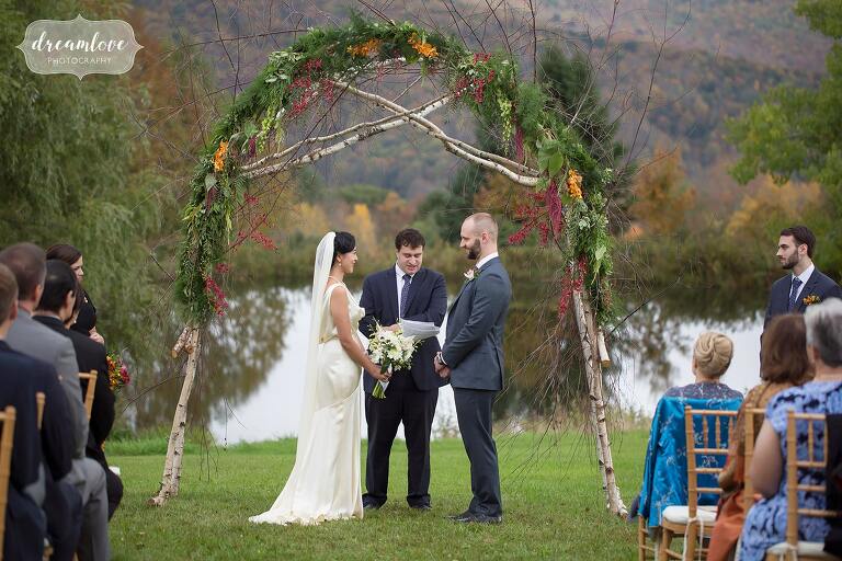 The bride and groom stand in front of their birchwood arbor with fall greenery in Roxbury, NY.