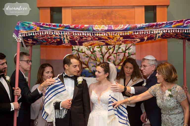 Family puts blanket around couple during Jewish wedding ceremony at Temple Shir Tikva.