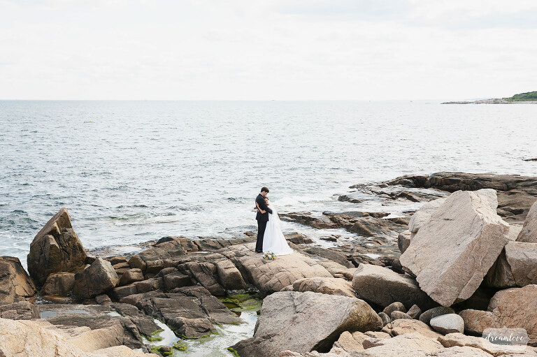 Bride and groom stand on rocks by ocean at Magnolia Beach in Gloucester, MA.