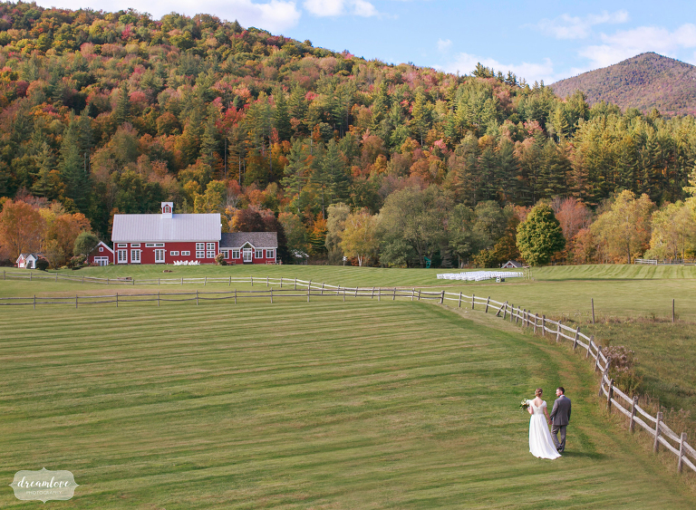 Bride and groom walk toward a red barn through a green field at Riverside Farm wedding venue in Vermont.